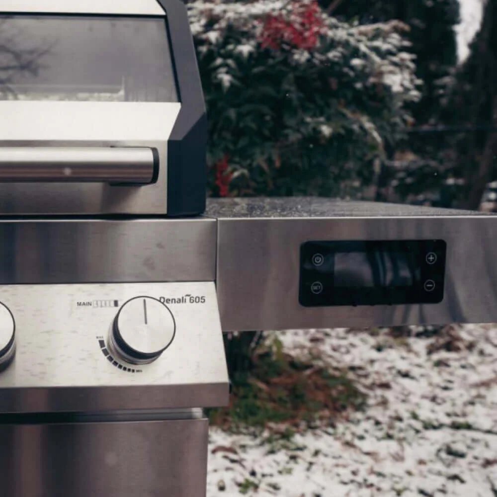 Stainless steel Monument Grills Denali 605 with control knobs in an outdoor setting with snow on the ground.