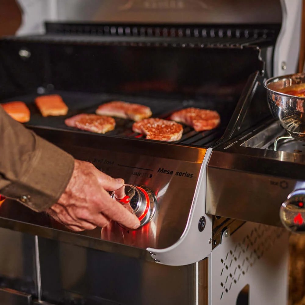 A person adjusting a gas Monument Grills Mesa 415BZ with food cooking on it