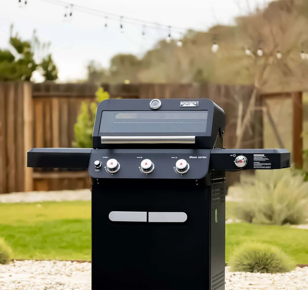 Black Monument Grills Mesa 325 in a backyard setting with greenery and a wooden fence.