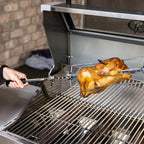 Person using a rotisserie to cook a chicken on a grill.