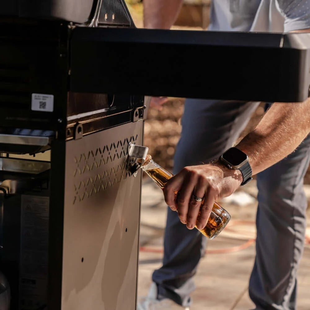 Person opening a barbecue grill with a bottle of beer