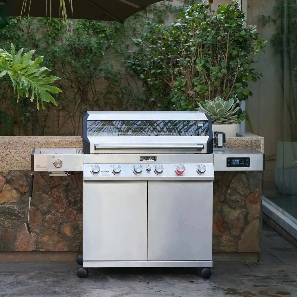 Stainless steel grill on wheels in an outdoor setting with plants and stone wall.