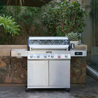 Stainless steel grill on wheels in an outdoor setting with plants and stone wall.
