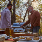 Two men grilling outdoors with a scenic background of trees and sunlight.