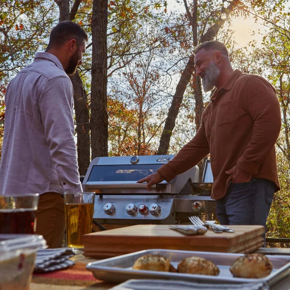 Two men grilling outdoors with a scenic background of trees and sunlight.