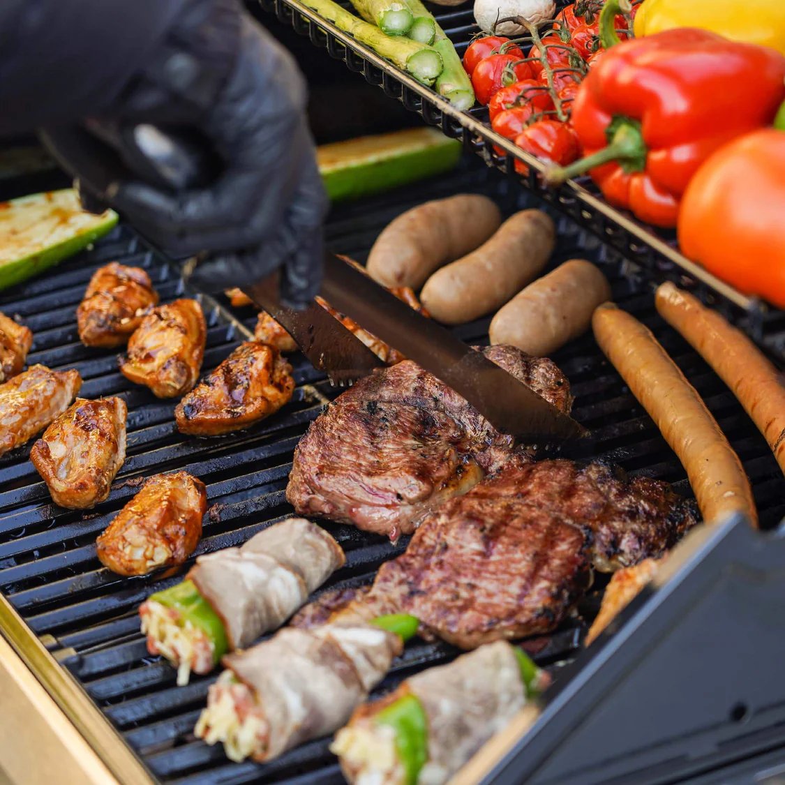 Grilling meat, vegetables, and sausages on a barbecue with a gloved hand using tongs.